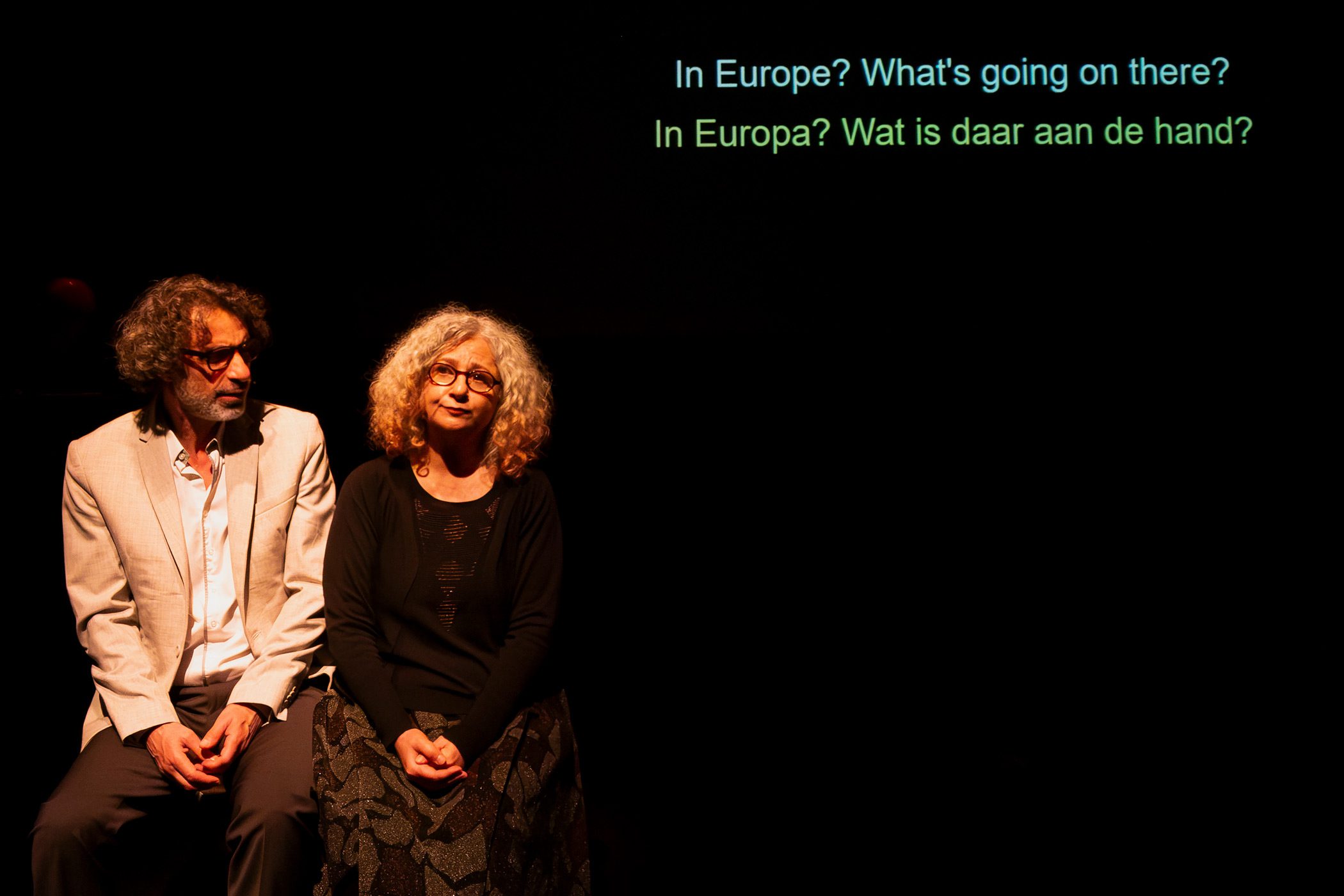 Zwei Personen sitzen auf einer Bank und sprechen miteinander. An der Wand im Hintergrund ist Schrift Projiziert.